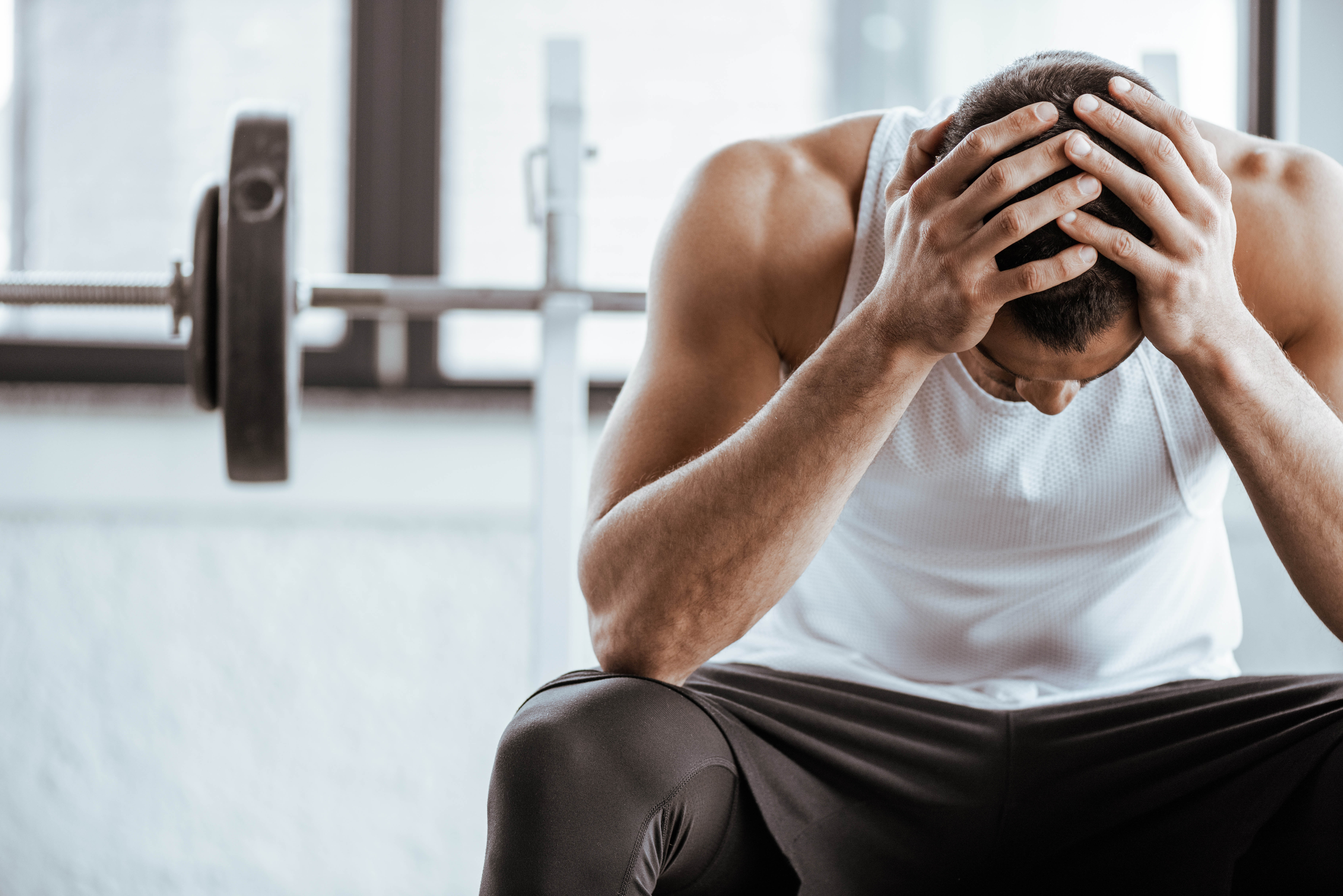 Fit young man sitting in the gym with his head in the hands, weights in the background